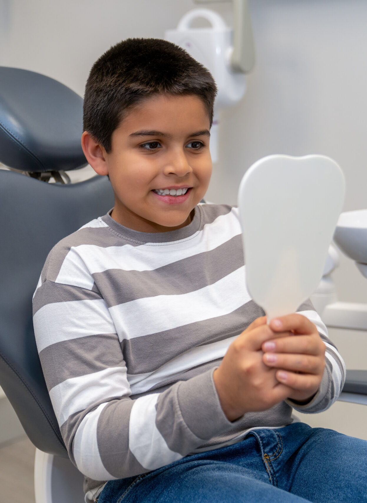 Happy boy sitting in dentist chair, holding a mirror and checking his teeth after dental checkup