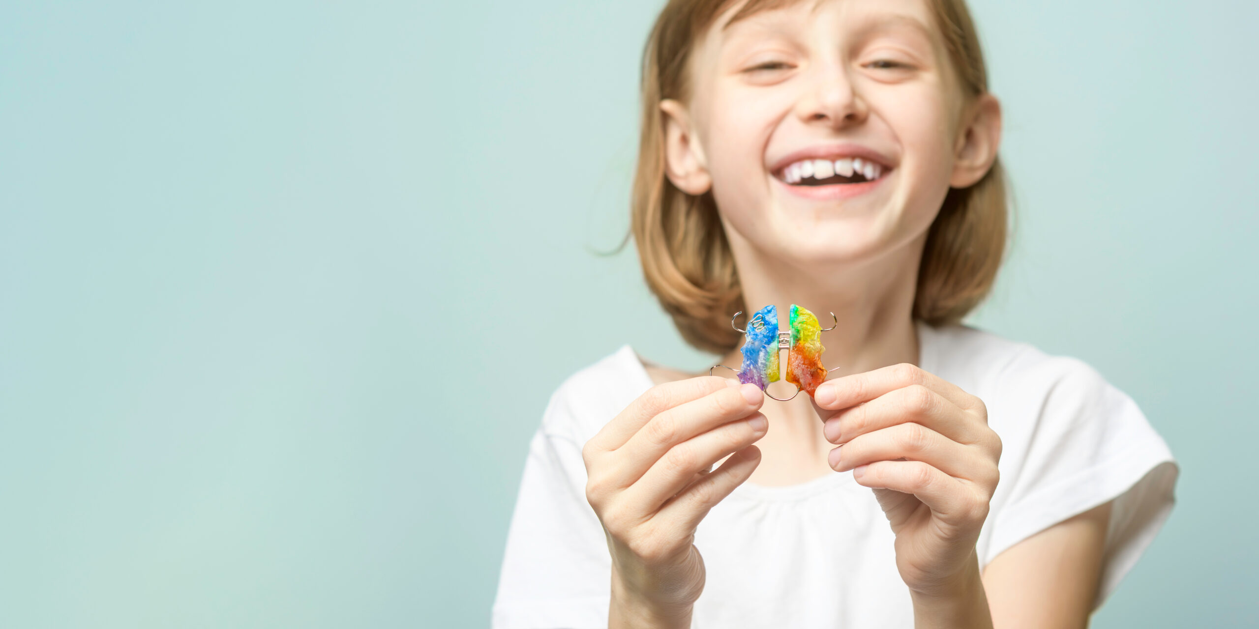 Smiling child holds rainbow orthodontic retainer on pastel background with copy space for dental clinic banner and kids care promotion.