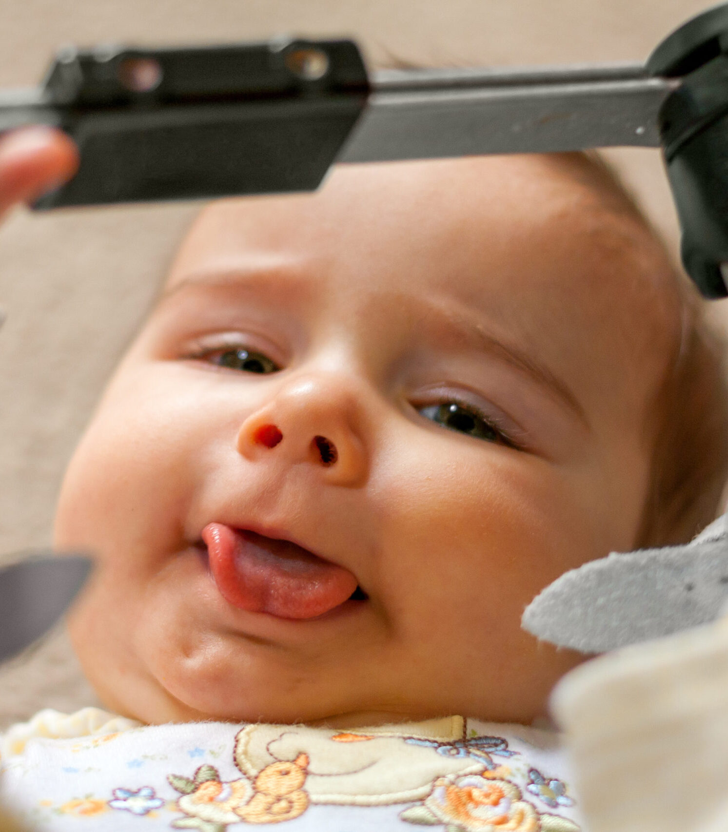 A nine month old baby with a tongue tie and orthopedic shoes lays on her back with her tongue sticking out as much as possible as she holds the bar between her orthopedic shoes that she wore for a club foot.