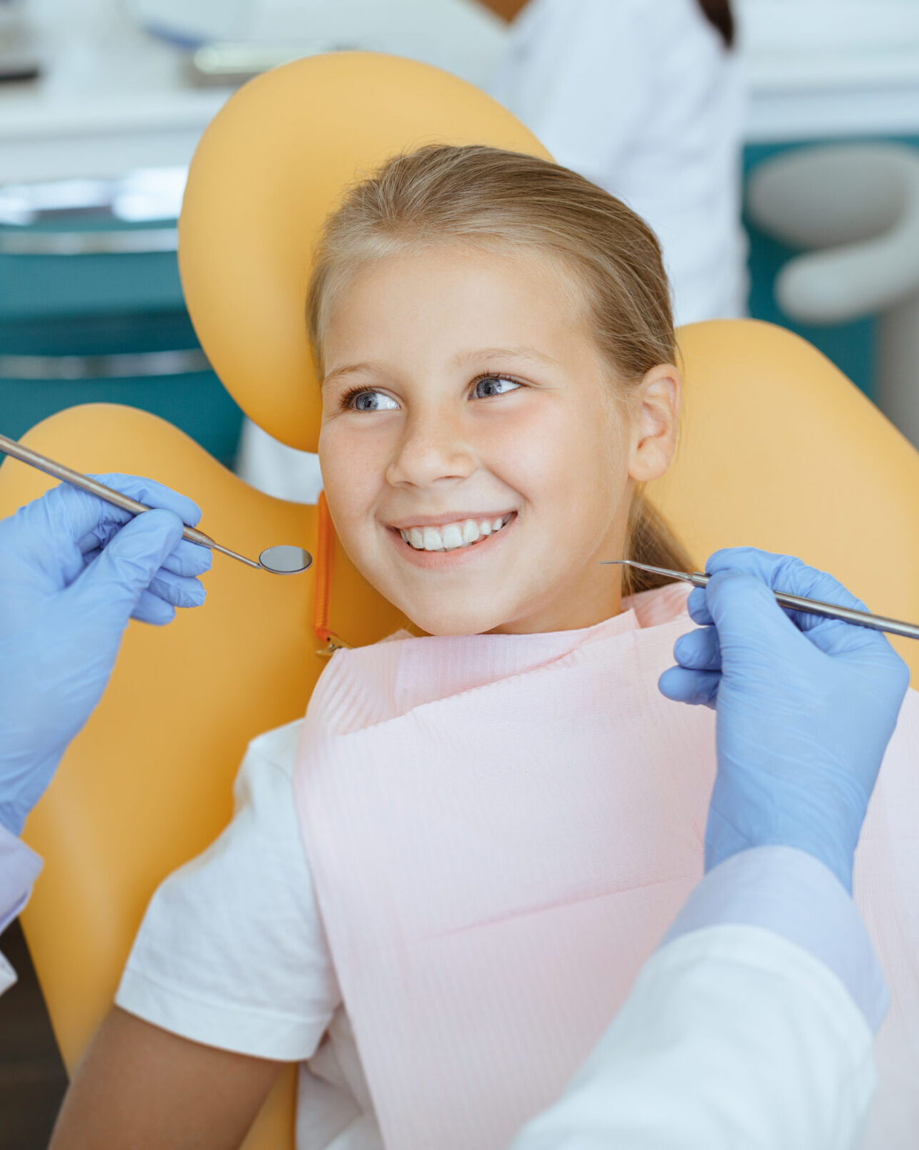 Fearless visit to doctor and dental treatment for child. Smiling girl in medical chair looks at dentist man in white coat, protective mask and rubber gloves with tools in his hands, and not afraid