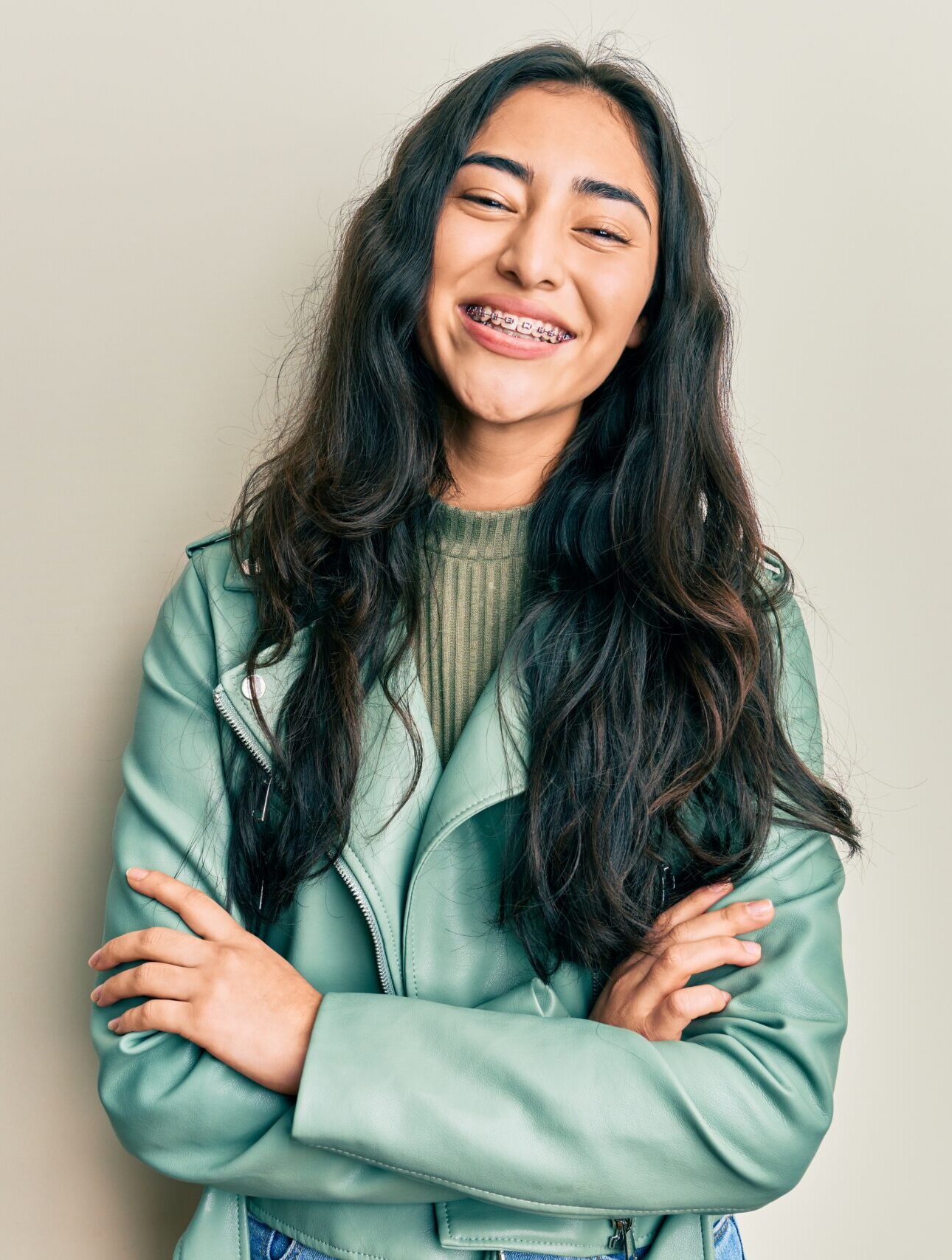 Hispanic teenager girl with dental braces wearing green leather jacket happy face smiling with crossed arms looking at the camera. positive person.