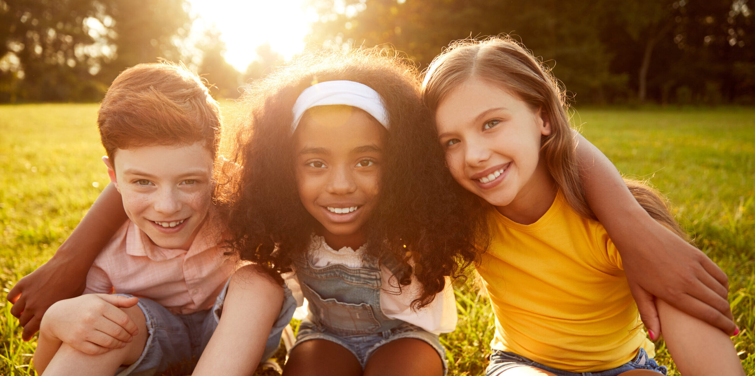 Cheerful black girl smiling for camera and embracing friends while sitting on lawn at sunrise in park